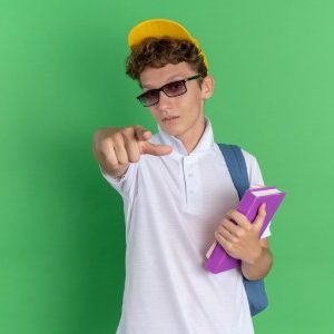 student-guy-white-shirt-yellow-cap-wearing-glasses-with-backpack-holding-notebooks-pointing-with-index-finger-camera-looking-confident (Custom)